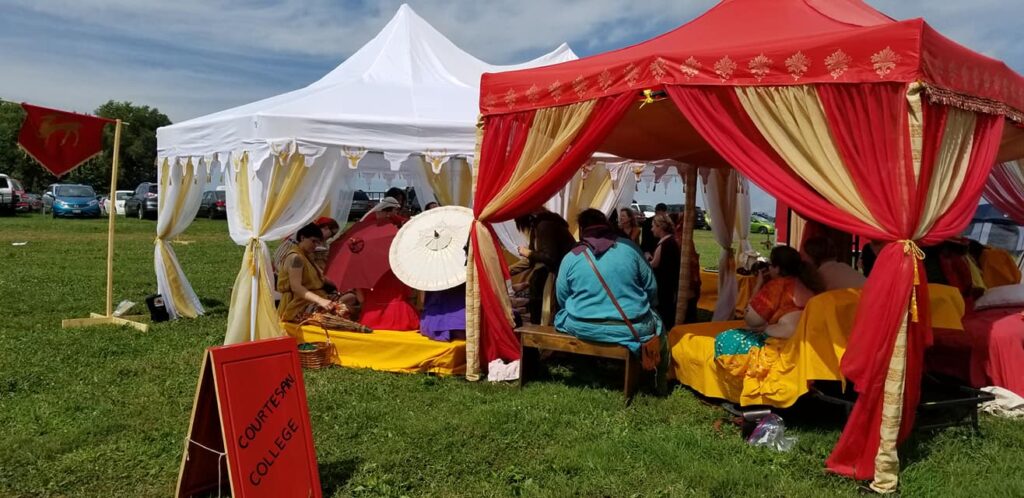 People in ancient Roman, medieval, and Renaissance clothing sitting and standing under a tent eating and talking
