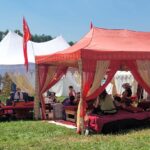 A group of people in ancient, medieval, and Renaissance clothing sitting under two fancy tents listening to one of them who is speaking