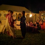 Guests at the first Pennsic Knowne World Courtesans Roman Bacchanal on August 7th, 2017 People in ancient Roman clothing standing and sitting while drinking, eating, and chatting outdoors at night