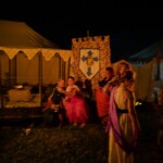 Guests at the first Pennsic Knowne World Courtesans Roman Bacchanal on August 7th, 2017 People in ancient Roman clothing standing and sitting while drinking, eating, and chatting outdoors at night