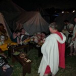 Guests at the first Pennsic Knowne World Courtesans Roman Bacchanal on August 7th, 2017 People in ancient Roman clothing standing and sitting while drinking, eating, and chatting outdoors at night