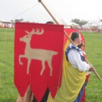 Courtesans prepare to process in Pennsic Opening Ceremonies on August 5, 2017
<br><br>
Photo by Godiva D'Mer A group of people in ancient Roman, medieval, and Renaissance clothing stand waiting in a field. One is carrying a red banner with a gold stag on it, which has one foreleg raised.
