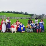 Courtesans, Patrons, and Tullians observing the Pennsic Unbelted Champions Battle on August 6, 2017 11 people dressed in ancient Roman, medieval, and Renaissance clothing, two of whom are wearing red veils, sitting and standing in a group photo on a grassy hill with a wooden fort in the background
