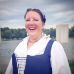 Courtesan Ciana Scholari di Polcenigo posing as she arrives at the Pennsic Knowne World Courtesans Social on August 10, 2017 A woman dressed in Renaissance clothing smiling with a lake in the background