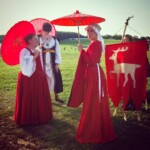 Courtesans Magdalena la Sanguigni and Esa inghean Donnchaidh prepare to process with Tullian Yenyega Velasconi de Santurce and another courtesan in Pennsic Opening Ceremonies on August 5th, 2017 4 women dressed in medieval and Renaissance clothing stand on a grassy hill, three carry red parasols and the fourth carries a red banner with a gold stag