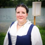 Courtesan Ciana Scholari di Polcenigo posing as she arrives at the Pennsic Knowne World Courtesans Social on August 10, 2017 A woman dressed in Renaissance clothing smiling with a lake in the background