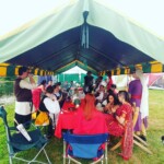 Courtesans eating and chatting around the table at the Pennsic Knowne World Courtesans Social on August 10, 2017 A group of people wearing ancient Roman, medieval, and Renaissance clothing sitting around a table under a large canopy eating and talking