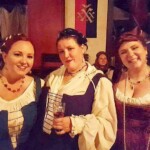 Courtesans Héloïse de Bruyères, Ciana Scholari di Polcenigo, and Amari bat Abraham at the Lowdy Toady at Casa Bardicci at Pennsic on August 6, 2017 3 women dressed in Renaissance clothing posing together with medieval style buildings in the background