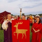 Courtesans Fortune St Keyne and Bianca Anguissola prepare to process with another courtesan in Pennsic Opening Ceremonies on August 5th, 2017 3 women dressed in ancient Roman clothing stand on a grassy hill with a crowd of reenactors in the background, one carries a red parasol and two support a red banner with a gold stag