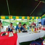 Courtesans eating and chatting around the table at the Pennsic Knowne World Courtesans Social on August 10, 2017 A group of people wearing ancient Roman, medieval, and Renaissance clothing sitting around a table under a large canopy eating and talking