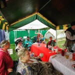 Courtesans eating and chatting around the table at the Pennsic Knowne World Courtesans Social on August 10, 2017 A group of people wearing ancient Roman, medieval, and Renaissance clothing sitting around a table under a large canopy eating and talking
