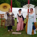 Courtesans Fortune St Keyne and Bianca Anguissola posing with friend Gritta of Strangewayes as they arrive at the Pennsic Knowne World Courtesans Social on August 10, 2017 Three courtesans wearing Renaissance and Persian clothing posing together, two holding parasols and a third carrying a closed parasol