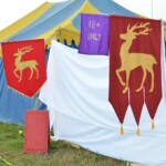 Setting up for the Pennsic Knowne World Courtesans Roman Bacchanal on August 6, 2018 Entrance to a camp featuring medieval tents, an "18+ only" banner, and two red banners with gold stags on them