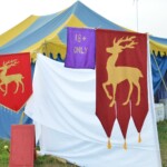 Setting up for the Pennsic Knowne World Courtesans Roman Bacchanal on August 6, 2018 Entrance to a camp featuring medieval tents, an "18+ only" banner, and two red banners with gold stags on them