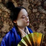 Courtesan Yamamura Kitsune at the Annual KWC Social at Pennsic in August of 2015 A person in Japanese clothing looking down and to the left holding a hand fan in front of an old fashioned map background