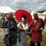 Courtesan Yamamura Kitsune at Pennsic in August of 2016
<br><br>
Photo by Heather Hall A person in Japanese clothing holding a red parasol standing between two people in medieval armor