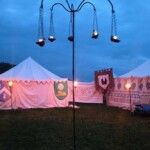 Replica ancient Roman lamps by <a class='blue-link' href="https://claybabypottery.com/">Claybaby Pottery<a/> hung on replica lampstands by Lucien de La Rochelle at the first Pennsic Knowne World Courtesans Roman Bacchanal on August 7th, 2017 A tall iron lampstand in the courtyard of a medieval camp at dusk