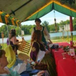 Courtesans Héloïse de Bruyères, Eadyth Woderose, Oriana of Xylina, and others drinking, eating, and chatting at the Pennsic Knowne World Courtesans Social on August 8th, 2016 People in ancient Roman, medieval, and Renaissance clothing gathered around a table under a tent