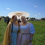 Courtesans Bianca Anguissola and Fortune St Keyne at Pennsic in August of 2015 Two women in Italian Renaissance clothing holding hand fans standing in a field surrounded by people in medieval clothing. One of the women is holding a parasol.