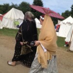 Courtesans Bianca Anguissola and Fortune St Keyne at Pennsic in August of 2015 Two women in Italian Renaissance clothing holding mugs walking on a dirt road by some medieval style tents. One is holding a parasol.