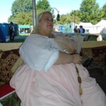 Courtesan Fortune St Keyne at Pennsic in August of 2015 A woman in Italian Renaissance clothing lounging on a couch holding a hand fan with picnic tables, a road, portable toilets, and medieval style tents in the background