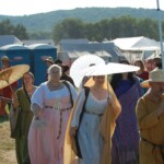 Courtesans Bianca Anguissola and Fortune St Keyne at Pennsic in August of 2015<br><br>Photo by Perronelle de Croy Two women in Italian Renaissance clothing holding hand fans walking on a dirt road surrounded by people in medieval clothing. One of the women is holding a parasol.