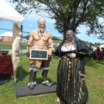 Courtesan Fortune St Keyne at Pennsic in August of 2015 A smiling woman in Italian Renaissance clothing holding a cup and using her thumb to indicate a life sized statue of a shirtless muscular man beside a medieval style tent