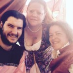 Courtesan Magdalena la Sanguigni, Tullian Yenyega Velasconi de Santurce, and friend Ezekiel at Pennsic on August 3, 2016 Closeup of three people in medieval and Renaissance clothing under a medieval style tent smiling at the camera