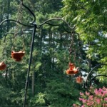 Replica ancient Roman lamps by <a class='blue-link' href="https://claybabypottery.com/">Claybaby Pottery<a/> hung on replica lampstands by Lucien de La Rochelle for the first Pennsic Knowne World Courtesans Roman Bacchanal in Atlantia on July 23, 2017 Two tall iron lampstands from which hang replica ancient Roman oil lamps on chains in a yard