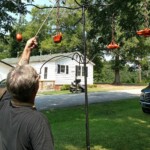 Replica ancient Roman lamps by <a class='blue-link' href="https://claybabypottery.com/">Claybaby Pottery<a/> hung on replica lampstands by Lucien de La Rochelle for the first Pennsic Knowne World Courtesans Roman Bacchanal in Atlantia on July 23, 2017 A person in modern clothing hanging a replica ancient Roman oil lamp on a chain on a tall iron lampstand in a yard