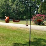 Replica ancient Roman lamps by <a class='blue-link' href="https://claybabypottery.com/">Claybaby Pottery<a/> hung on replica lampstands by Lucien de La Rochelle for the first Pennsic Knowne World Courtesans Roman Bacchanal in Atlantia on July 23, 2017 Two tall iron lampstands from which hang replica ancient Roman oil lamps on chains in a yard