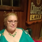 Courtesan Fortune St Keyne at Pennsic on August 4, 2017 A woman in ancient Roman clothing and a gold stag pin sitting on a bench next to a sign that reads "Cooper's Lake"