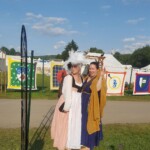 Courtesan Bianca Anguissola with friend Gritta of Strangewayes at Pennsic on August 5, 2017 A person in German Renaissance clothing standing next to a woman in ancient Roman clothing who is waving in front of some decorated fabric forming walls outdoors