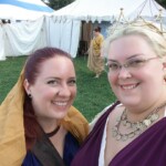 Courtesans Bianca Anguissola and Fortune St Keyne at Pennsic on August 5, 2017 Closeup of two smiling women wearing ancient Roman clothing in front of several medieval style tents with another person in ancient Roman clothing walking in the background