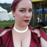 Courtesan Bianca Anguissola before the first Pennsic Knowne World Courtesans Roman Bacchanal on August 7th, 2017 Closeup of a woman in ancient Roman clothing wearing a gold stag pin with medieval style tents in the background