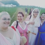 Courtesans Bianca Anguissola and Fortune St Keyne with patron Keziah Planchet and friend Simona bat Leone at Pennsic on August 9, 2017 Four people in ancient Roman and medieval clothing posing for a photo with medieval style tents, a field, a dirt road, and other people dressed in ancient and medieval clothing behind them