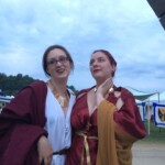Courtesan Bianca Anguissola with friend Gritta of Strangewayes before the first Pennsic Knowne World Courtesans Roman Bacchanal on August 7th, 2017 Two people in ancient Roman clothing, one of whom is wearing a gold stag pin, standing together and posing for a photo outdoors at dusk