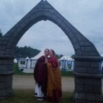Courtesan Bianca Anguissola with friend Gritta of Strangewayes before the first Pennsic Knowne World Courtesans Roman Bacchanal on August 7th, 2017 Two people in ancient Roman clothing, one of whom is wearing a gold stag pin, standing with their palms touching and posing for a photo under a large faux stone arch outdoors at dusk