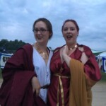 Courtesan Bianca Anguissola with friend Gritta of Strangewayes before the first Pennsic Knowne World Courtesans Roman Bacchanal on August 7th, 2017 Two people in ancient Roman clothing, one of whom is wearing a gold stag pin, standing together and posing for a photo outdoors at dusk