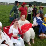 Courtesans, patrons, Tullians, and friends observing the Pennsic Unbelted Champions Battle on August 6th, 2017 A group of people in ancient, medieval, and Renaissance clothing, two of whom are wearing red veils, sitting and standing in a field.