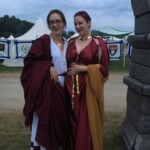 Courtesan Bianca Anguissola with friend Gritta of Strangewayes before the first Pennsic Knowne World Courtesans Roman Bacchanal on August 7th, 2017 Two people in ancient Roman clothing, one of whom is wearing a gold stag pin, standing together and posing for a photo outdoors at dusk