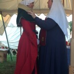 Courtesan Esa inghean Donnchaidh with her Laurel Beatriz Aluares de la Oya at their apprentice ceremony at Pennsic on August 3, 2017 A person in medieval clothing adjusting the hood of another person in medieval clothing inside a medieval style tent