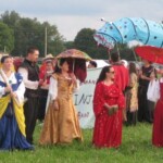 Courtesans prepare to process in Pennsic Opening Ceremonies on August 5, 2017 <br><br>Photo by Godiva D'Mer A group of people in ancient Roman, medieval, and Renaissance clothing stand waiting in a field. One is carrying a red banner with a gold stag on it, which has one foreleg raised. Another is holding a red parasol.