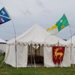Courtesan Magdalena la Sanguigni and Tullian Yenyega Velasconi de Santurce's tent with the Hart of Gold banner at Pennsic in August of 2018 A medieval style tent with a red banner with a gold stag with one foreleg raised on it leaning against the side