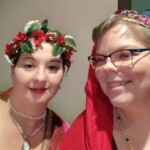 Courtesans Honora de Cunningham and Henriette Osanne Torchier at Twelfth Night in Northshield on January 14, 2023. Closeup of two people in Renaissance clothing, one of whom is wearing a red veil, smiling at the camera indoors