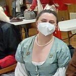 Courtesan Ginevra Fiammetta di Silvestri at Feast of St. Sebastian in Atlantia on February 21st, 2023.<br><br>Photo by Zofia Anzelmanka A woman in Renaissance clothing wearing a red veil, a gold stag pin, and a surgical mask sitting and smiling at the camera indoors