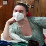 Courtesan Ginevra Fiammetta di Silvestri at Feast of St. Sebastian in Atlantia on February 21st, 2023.<br><br>Photo by Zofia Anzelmanka A woman in Renaissance clothing wearing a red veil, a gold stag pin, and a surgical mask sitting while leaning on a table and smiling at the camera indoors