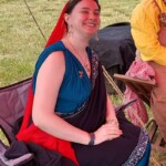 Courtesan Ginevra Fiammetta di Silvestri at Ruby Joust in Atlantia on May 27th, 2023<br><br>Photo by Zofia Anzelmanka A smiling woman in ancient Roman clothing wearing a red veil and a gold stag pin sitting in a modern camp chair outdoors