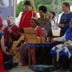 Courtesan Ginevra Fiammetta di Silvestri at Ruby Joust in Atlantia on May 27th, 2023
<br><br>Photo by Tannis of Tir-y-Don A woman in ancient Roman clothing wearing a red veil kneeling in front of two people in medieval clothing wearing crowns sitting on thrones with other people in medieval clothing standing behind them