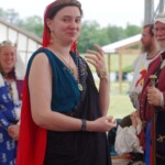 Courtesan Ginevra Fiammetta di Silvestri at Ruby Joust in Atlantia on May 27th, 2023
<br><br>Photo by Tannis of Tir-y-Don A woman in ancient Roman clothing wearing a red veil and a medallion standing in front of other people in medieval clothing